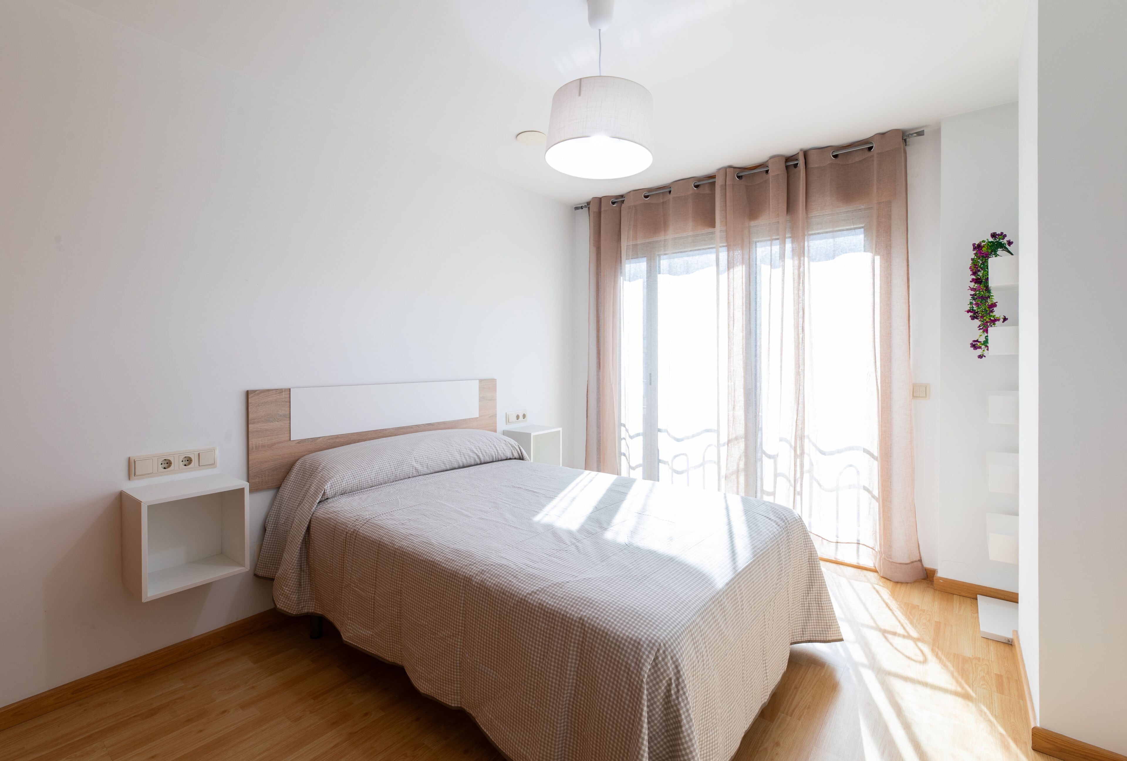 Un dormitorio con cama, una mesita de noche, una lámpara colgante, una ventana con cortinas y una planta pequeña en la pared.
