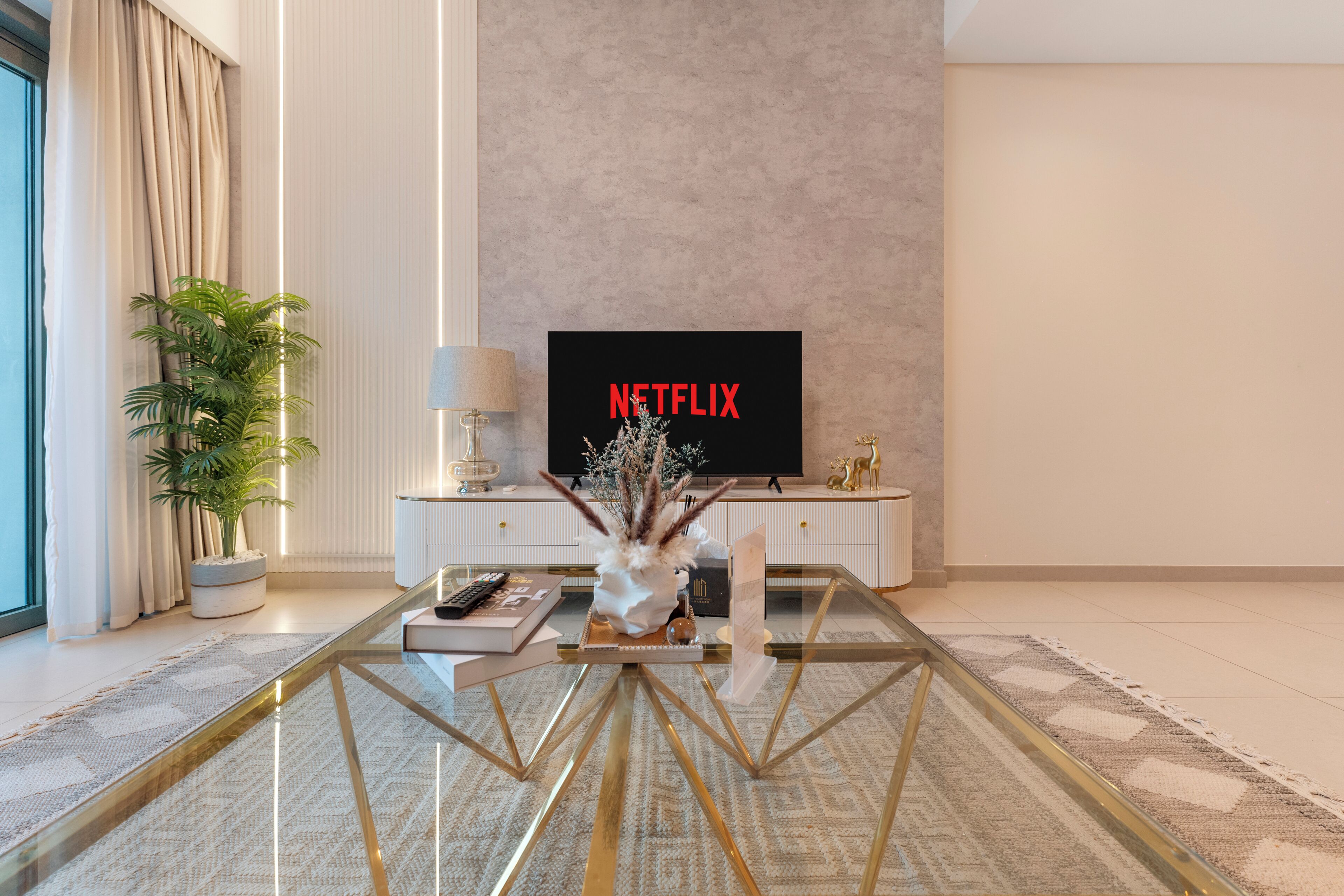 A modern living room with a glass coffee table, a television displaying 'NETFLIX,' a potted plant, and a decorative vase.
