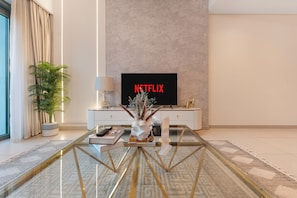 A modern living room with a glass coffee table, a television displaying 'NETFLIX,' a potted plant, and a decorative vase. - Walk to Dubai Mall and Dubai Fountain Parking Pool (Dubai)