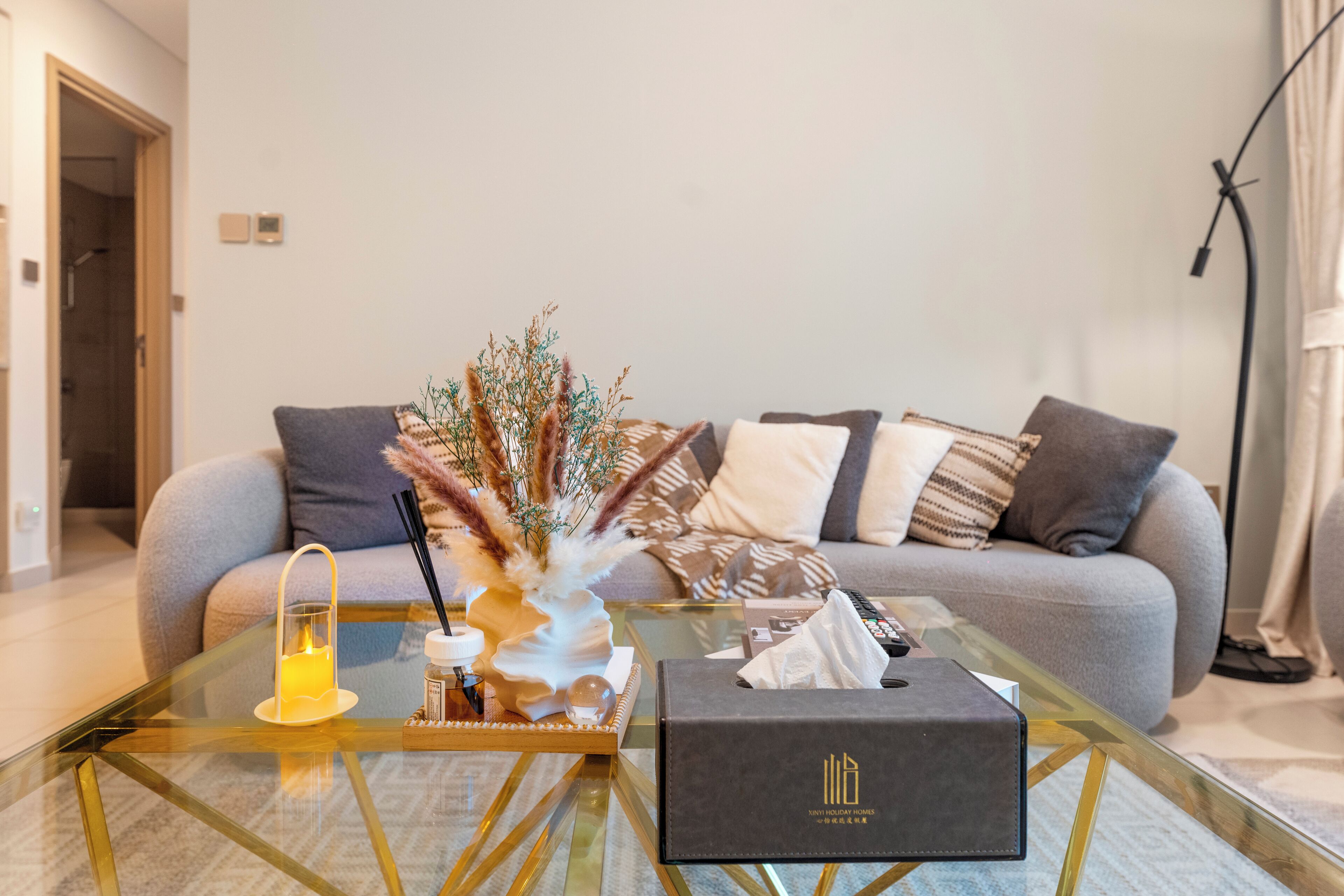 A modern living room with a glass coffee table, a sofa with patterned cushions, and a decorative vase with dried plants.