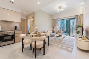Eine moderne Küche mit Einbauherd, ein Essbereich mit rundem Tisch und Stühlen sowie ein Wohnzimmer mit Sofa und Blick auf hohe Gebäude.