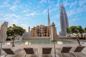 A poolside area with lounge chairs and umbrellas, a building with a unique architectural design, and a tall skyscraper in the background. - Walk to Dubai Mall and Dubai Fountain Parking Pool (Dubai)
