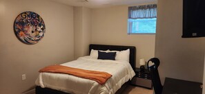 Une chambre à coucher comprenant un lit, une table de chevet, une lampe, une fenêtre avec des rideaux et une œuvre d’art murale.