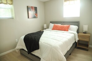 Ein ordentlich bezogenes Bett mit einer schwarz-weißen Decke, einem einzelnen orangefarbenen Kissen und einem grauen Kopfteil. An jeder Seite des Bettes steht ein Nachttisch mit einer Lampe.
