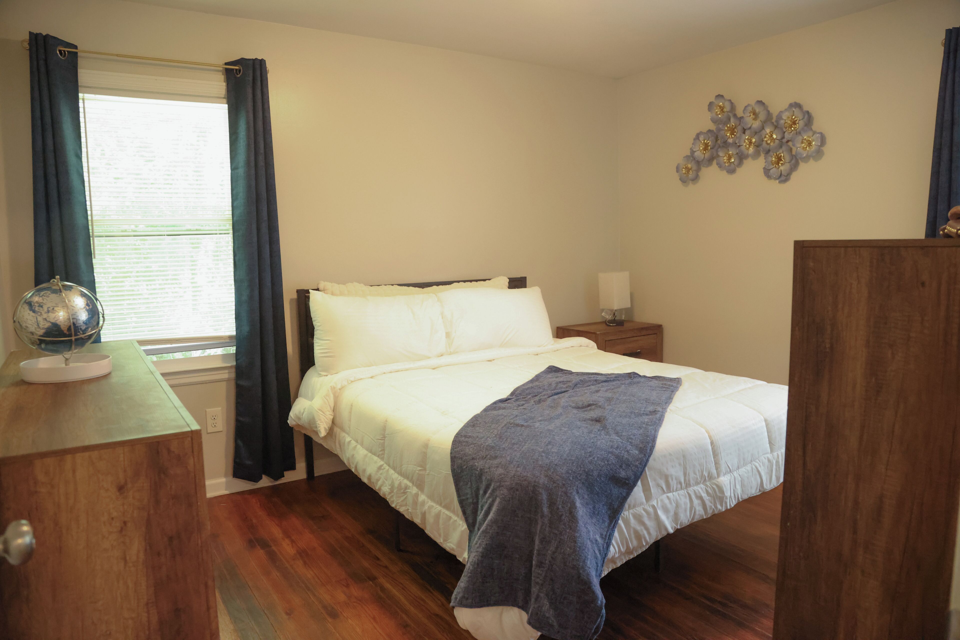 A bedroom with a bed, a nightstand, a dresser, a globe, and a window with curtains.