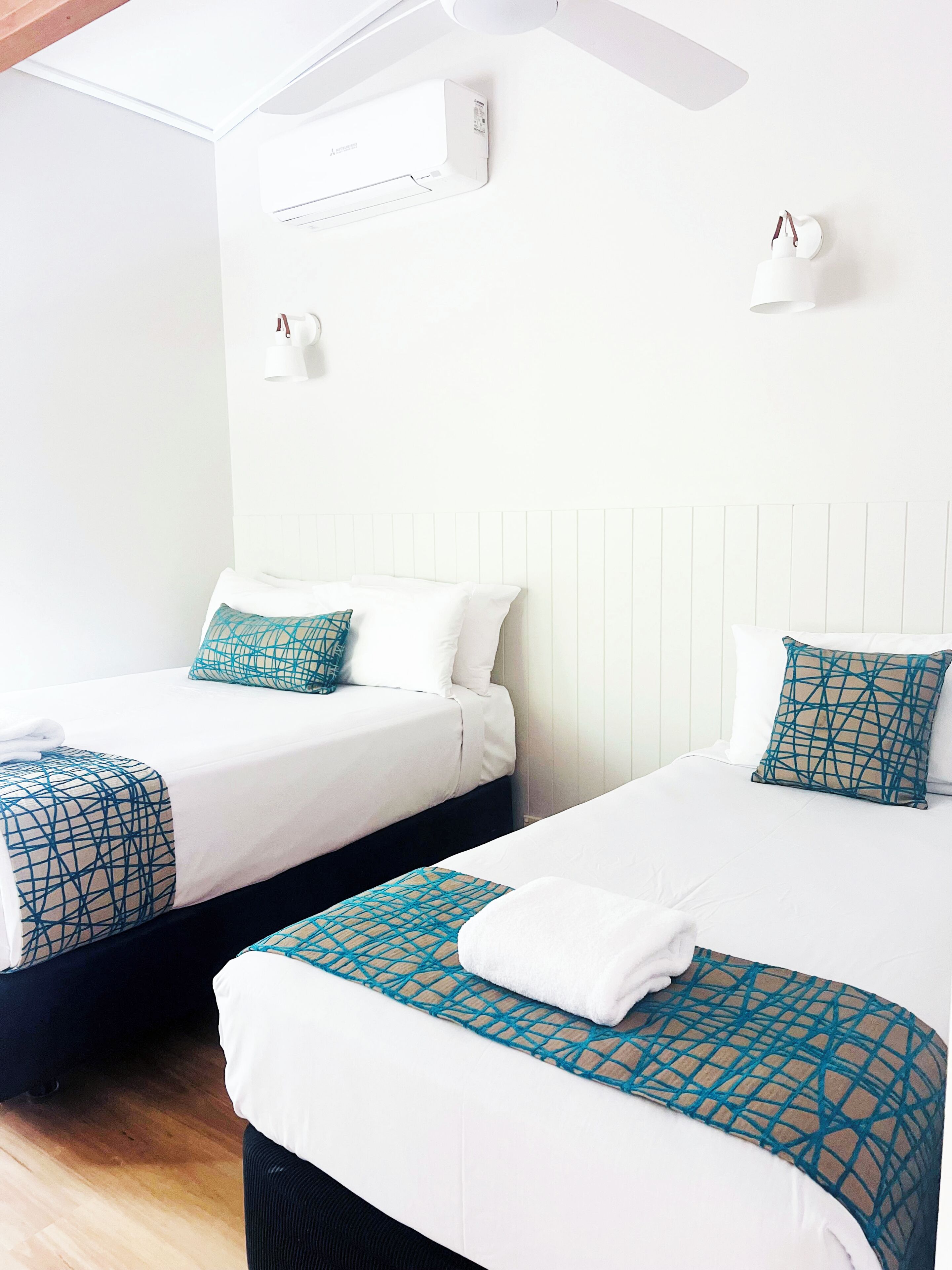 Two single beds with white bedding and teal geometric-patterned throw pillows, a white ceiling fan, and a wall-mounted air conditioning unit.