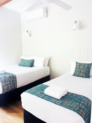 Duas camas de solteiro com roupa de cama branca e almofadas decorativas geométricas em tom verde-água, um ventilador de teto branco e um ar condicionado fixado na parede.