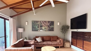 A spacious living room with a large sofa, a round coffee table, and a ceiling fan. There is a television mounted on the wall and a window providing natural light.