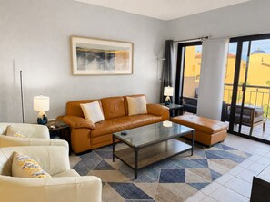 A modern living room with a sofa, armchair, and ottoman, a glass coffee table, and a large window with a view of buildings. - 2BDR Remodeled penthouse / Sandy Beach+ beach gear (Puerto Peñasco)