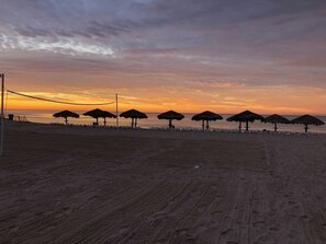 Ein Strand mit Sonnenschirmen und Liegestühlen, einem Volleyballnetz und einem Sonnenuntergang.