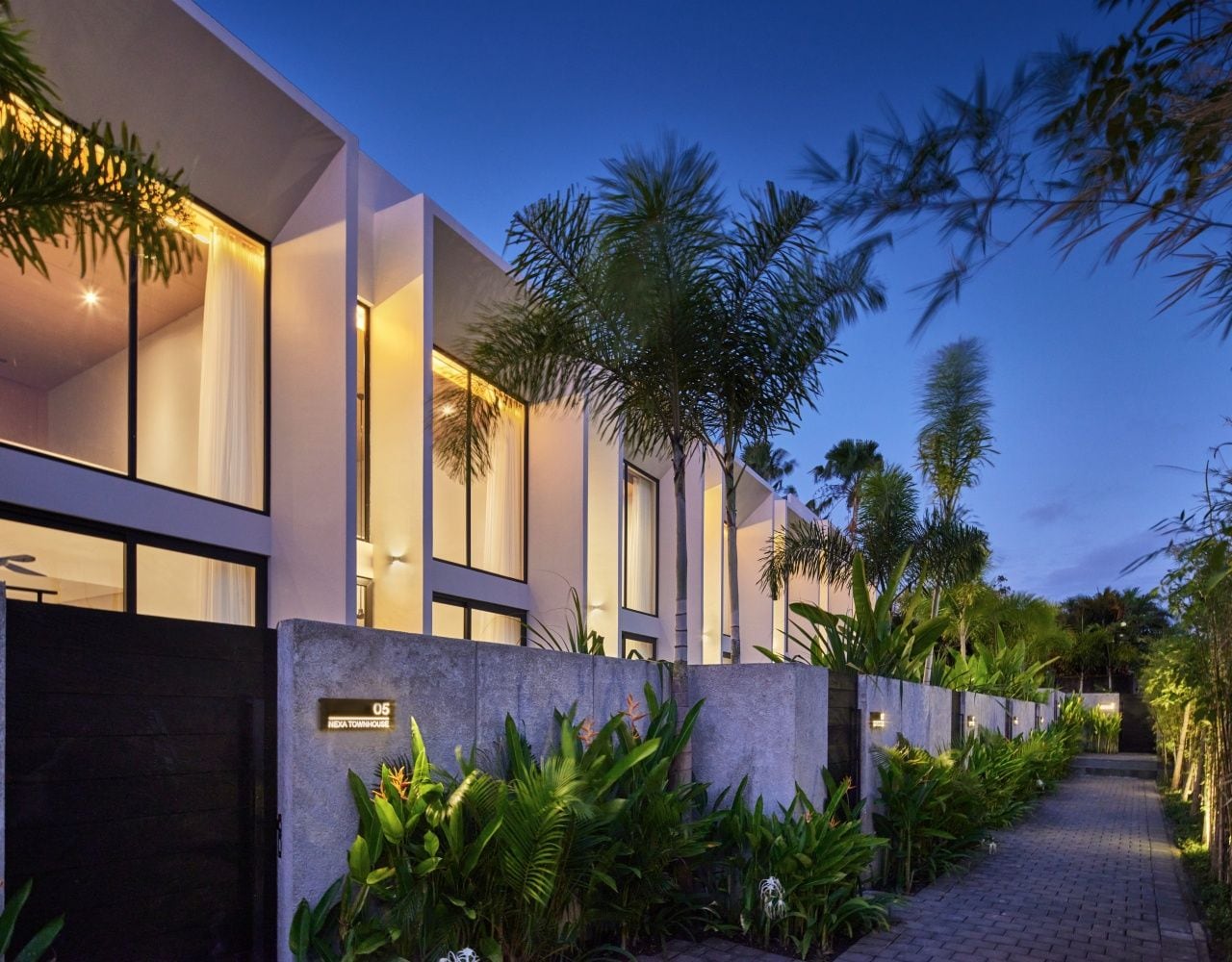 A modern building with a pathway lined with tropical plants, a dark entrance gate, and illuminated windows.