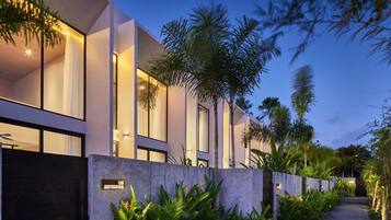 A modern building with a pathway lined with tropical plants, a dark entrance gate, and illuminated windows.