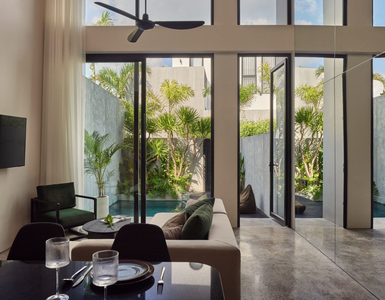 A modern living room with a glass door leading to a pool area, a ceiling fan, and a dining table set with glasses and cutlery.