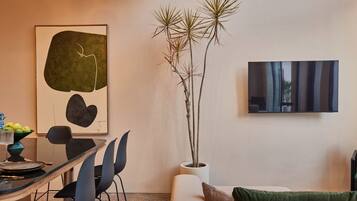 A modern living room with a dining area, featuring a glass table, black chairs, a potted plant, and artwork on the walls.