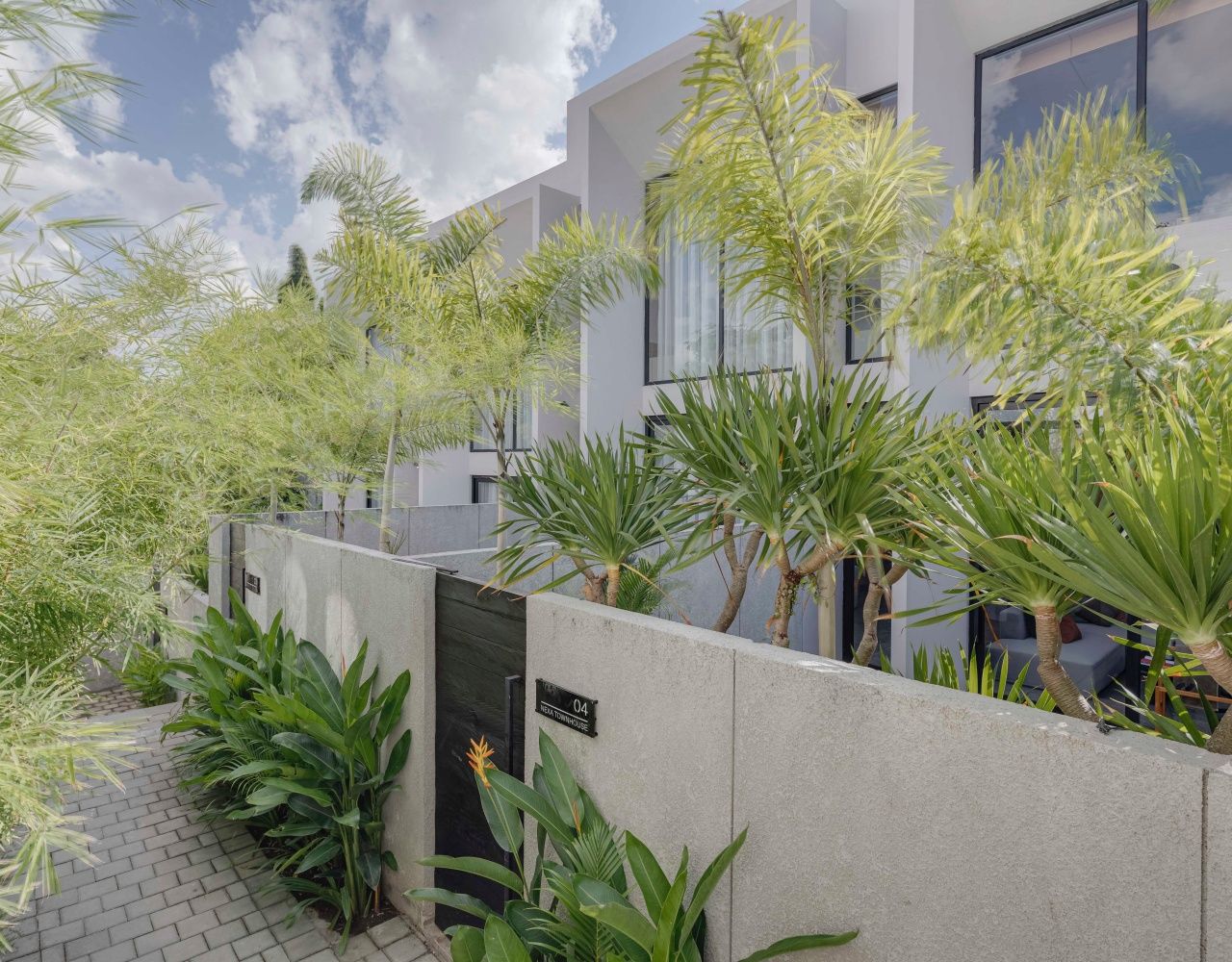 Un edificio moderno con fachada de hormigón, acceso pavimentado y abundante vegetación, incluyendo palmeras y otras plantas.