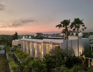 Un edificio moderno con numerosas ventanas y techo plano, rodeado de vegetación y palmeras.