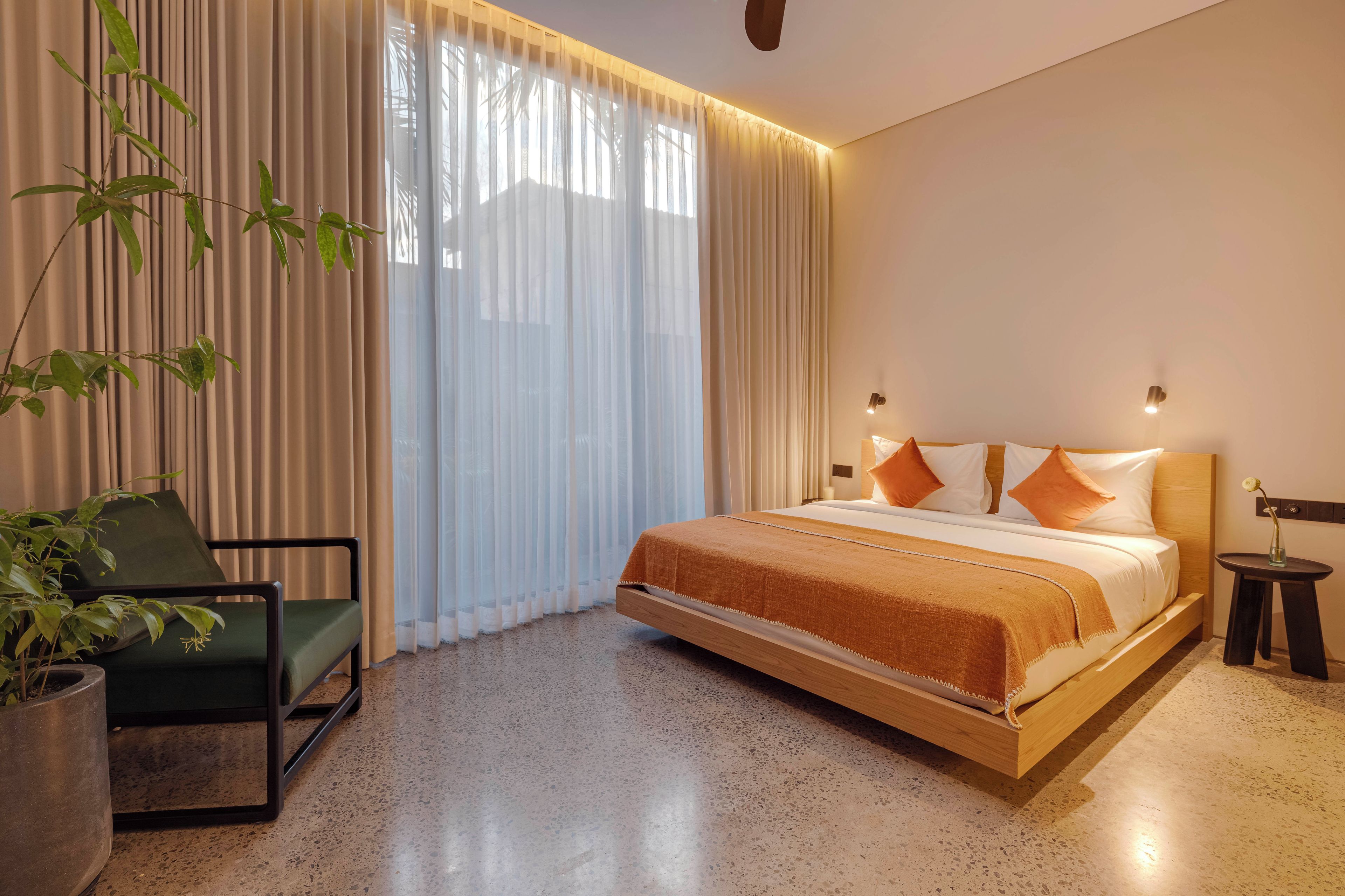 A modern hotel room with a bed, a chair, a potted plant, and a nightstand.