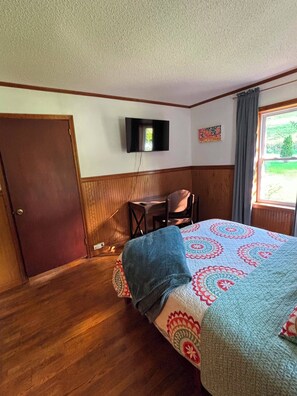 Una habitación con cama, escritorio, silla, televisión en la pared y ventana.