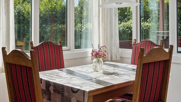 Uma mesa de jantar com toalha estampada, rodeada por cadeiras com estofamento listrado, diante de grandes janelas com vista para o verde.