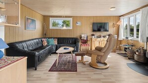 Uma sala de estar mobiliada com um sofá de couro preto, uma mesa de centro de madeira, um tapete estampado, uma cadeira de balanço de madeira e uma televisão montada na parede.