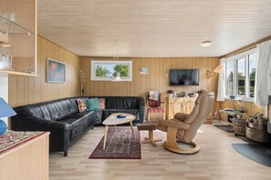 Ein möbliertes Wohnzimmer mit einer schwarzen Ledersofa, einem Holztisch, einem gemusterten Teppich, einem Holzschaukelstuhl und einem an der Wand montierten Fernseher.
