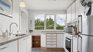 Uma cozinha moderna com armários brancos, uma geladeira de aço inoxidável e uma janela com vista para árvores e céu.