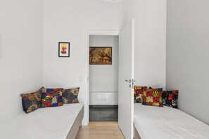 Ein kleiner, gut beleuchteter Raum mit zwei Betten, jedes mit bunten Kissenbezügen. Eine Tür führt ins Badezimmer, und ein gerahmtes Bild hängt an der Wand.