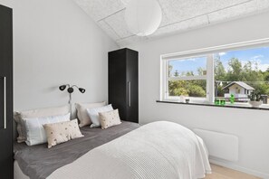 Ein Schlafzimmer mit einem Bett, Nachttischlampen, einem Fenster mit Blick auf Grünflächen und ein Haus, sowie einer weißen Decke.