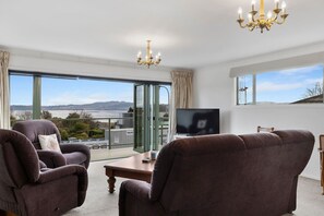 A modern living room with a television, two armchairs, a coffee table, and a view of the city through large windows. - Family Haven, Lake Taupō Escape (Taupō)