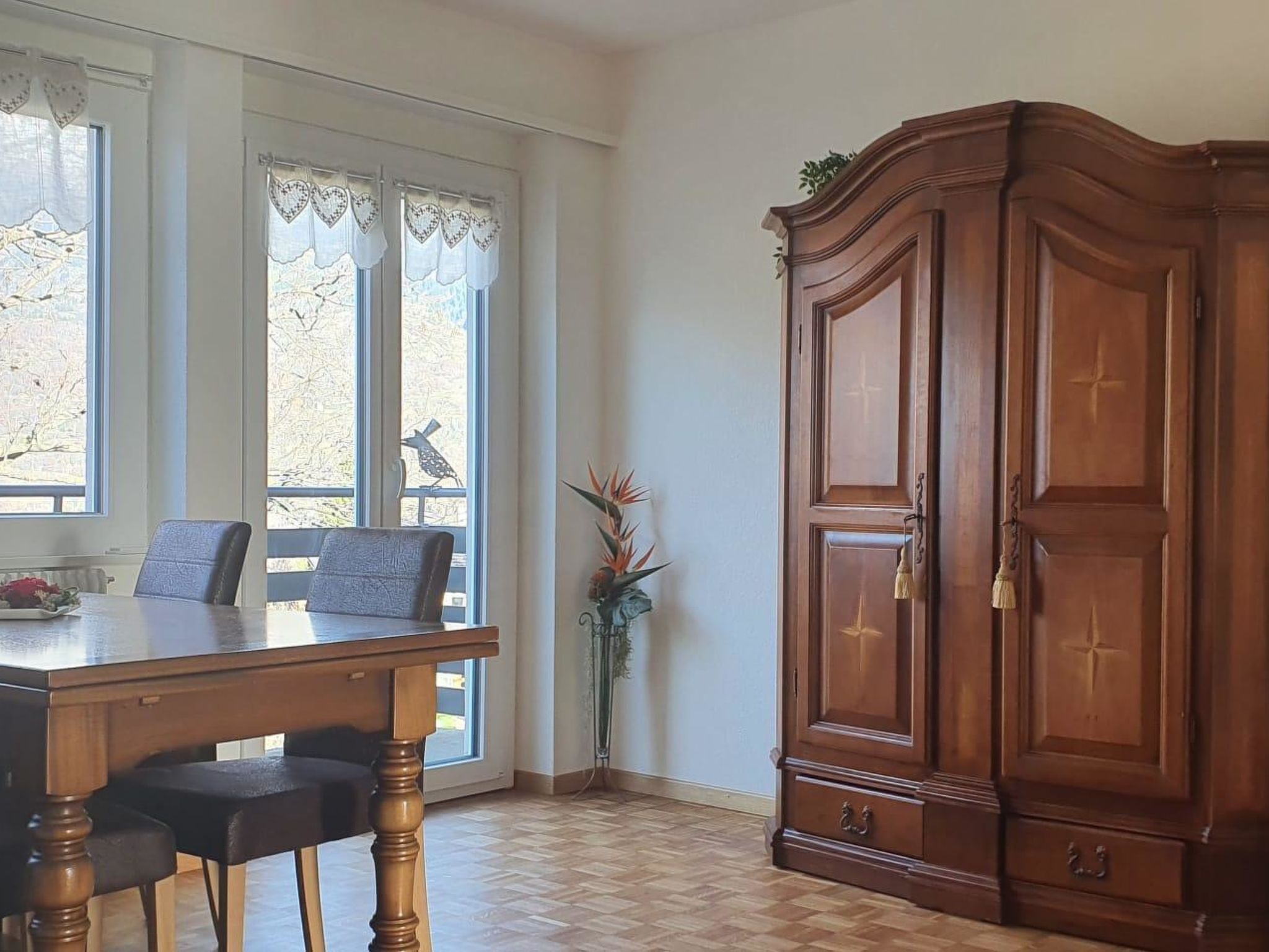 Une pièce avec une table en bois, des chaises, une grande armoire en bois et une fenêtre avec des rideaux.