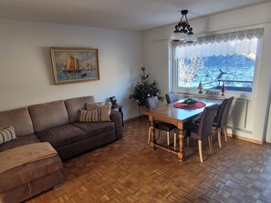 Ein Wohnzimmer mit braunem Sofa, einem Esstisch mit Stühlen, einer kleinen Weihnachtstanne und einem Gemälde an der Wand.