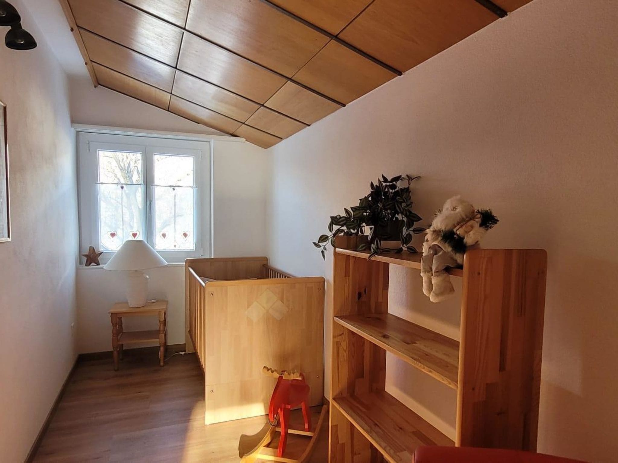 Quarto com berço de madeira, uma mesinha, prateleira com planta e um bicho de pelúcia, e janela com enfeite de estrela.