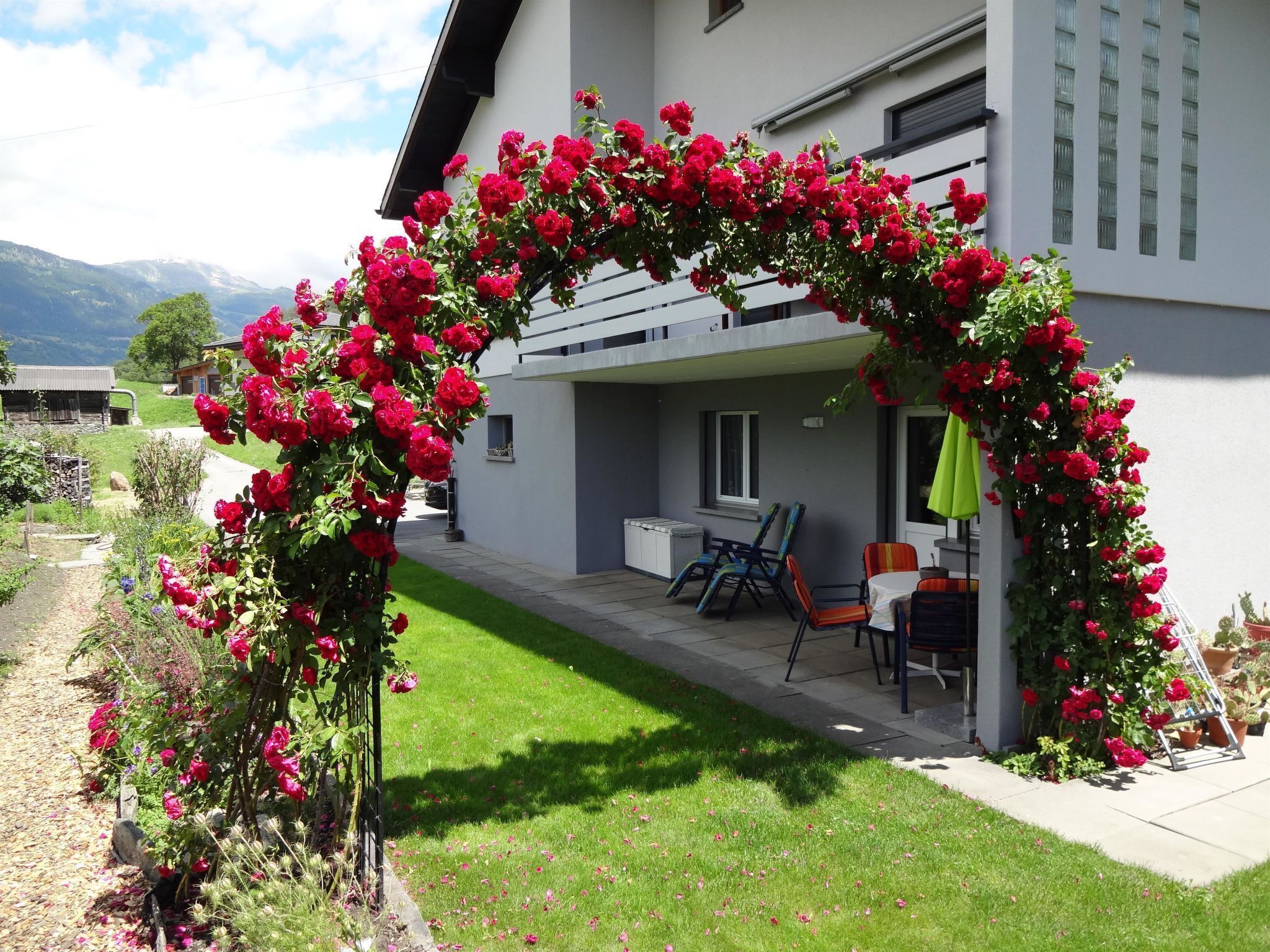 A modern house with a covered patio area, a small outdoor seating arrangement, and a garden with a flower arch.