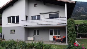 Ein zweistöckiges Gebäude mit einem Balkon, einem kleinen Garten und einer Bergkulisse.