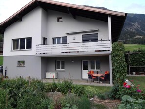 Ein zweistöckiges Gebäude mit einem Balkon, einem kleinen Garten und einer Bergkulisse.