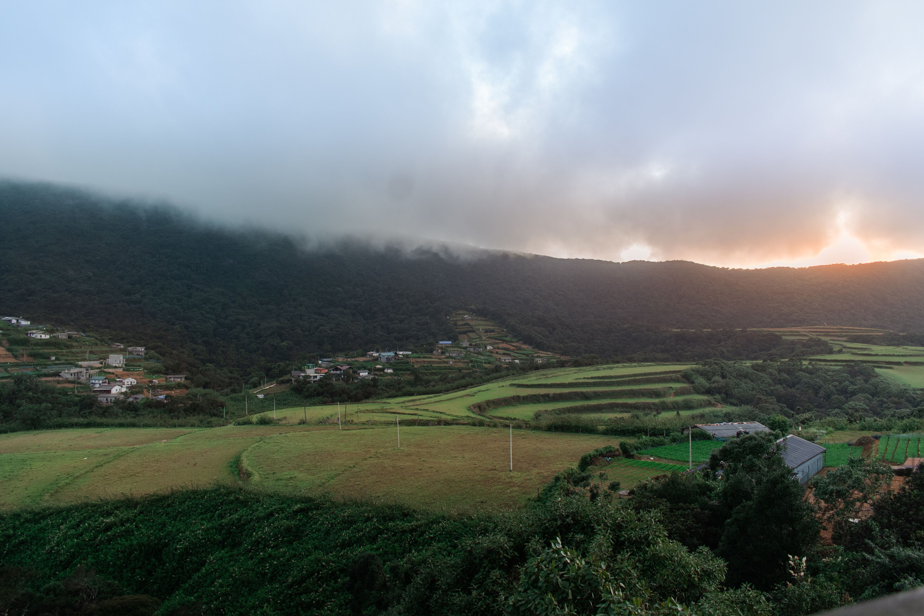 A rural landscape with terraced fields, scattered houses, and a mountain range in the background.