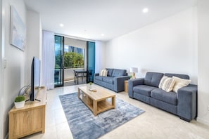 Een moderne woonkamer met een flatscreen televisie, twee banken, een salontafel en een balkon met uitzicht op groen en gebouwen.