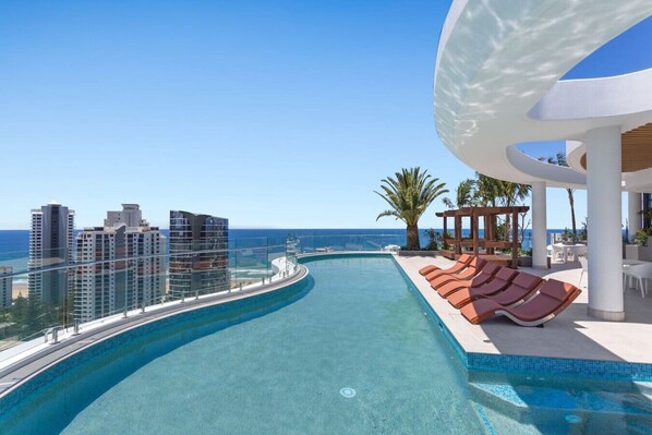 Una piscina de borde curvo, sillones de descanso y vista al océano y edificios.