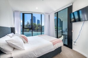 Habitación de hotel moderna con una cama grande, un televisor montado en la pared y un ventanal corredero que da a un balcón con vistas a la ciudad.