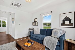 Ein modernes Wohnzimmer mit einem blauen Sofa, einem Couchtisch mit einem Buch und einem großen Fenster mit Blick auf Grünflächen.