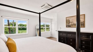 A four-poster bed with a white bedspread and a mustard-colored pillow, a dark wooden dresser with multiple drawers, and a large window with a view of greenery. - Iconic urban Bourbon Trail suite with EV charger! (Louisville)
