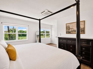 Ein Bett mit Baldachin, weiße Bettdecke und ein senffarbenes Kissen, eine dunkelholzausgekleidete Kommode mit mehreren Schubladen und ein großes Fenster mit Blick auf Grünflächen.