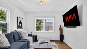 A modern living room with a grey sofa, a coffee table, a TV mounted on the wall, and a window with a view of greenery. - Iconic urban Bourbon Trail suite with EV charger! (Louisville)