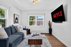 Ein modernes Wohnzimmer mit grauem Sofa, Couchtisch, Wandfernseher und einem Fenster mit Blick auf Grünflächen.