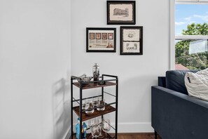 Ein Zimmer mit einem dunkelblauen Sofa, einem Metallregal mit Gläsern und einer Flasche sowie drei gerahmten Bildern an der Wand.