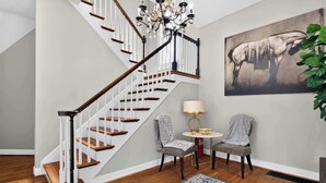 Un escalier avec des marches en bois et des rampes blanches, un lustre, deux chaises, une table avec une lampe et un tableau représentant un cheval accroché au mur.