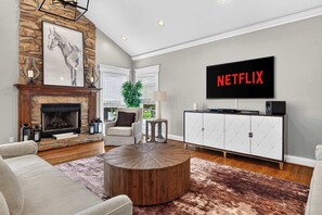 Ein Wohnzimmer mit Kamin aus Stein, einem runden Couchtisch, einer Couch, einem an der Wand befestigten Fernseher und einer Kommode mit Netflix-Logo.