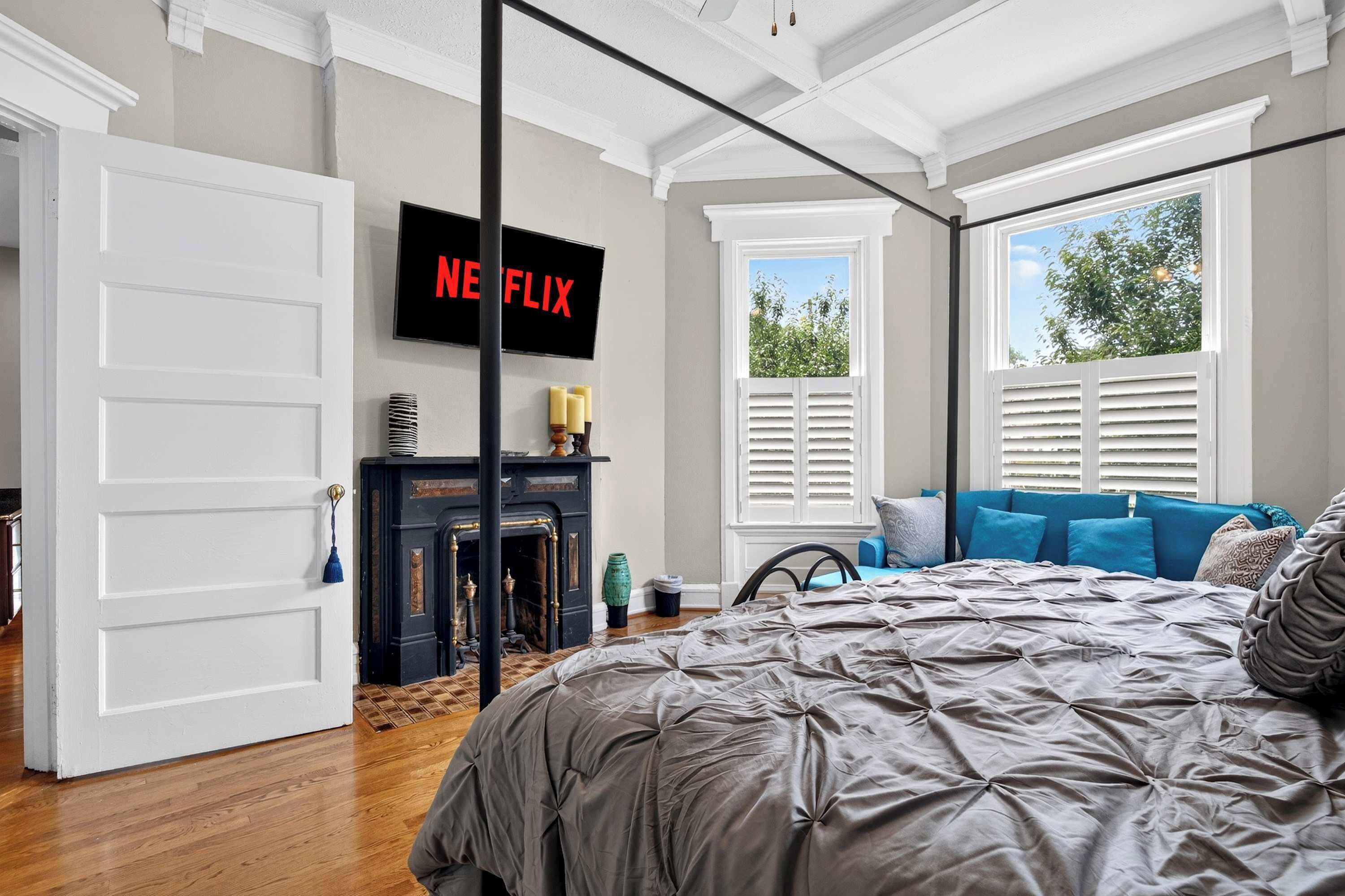 A bedroom with a large bed, a fireplace, a television displaying 'Netflix', and a window with blinds.