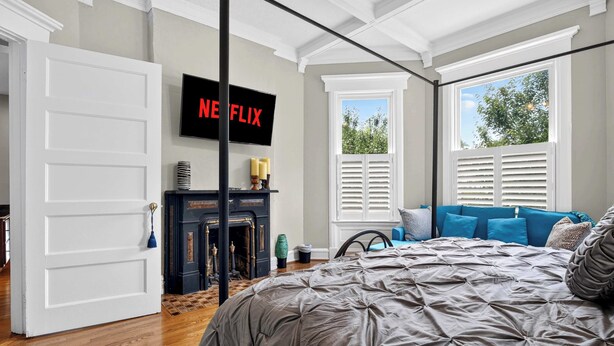Une chambre à coucher avec un grand lit, une cheminée, un téléviseur diffusant du contenu de « Netflix » et une fenêtre avec des stores.