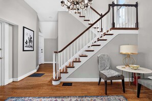 A staircase with wooden steps and white railings, a chandelier, a framed picture on the wall, a chair, a table with a lamp, and a rug on the floor. - 4-Bedroom Louisville Apartment | Backyard, Office (Louisville)